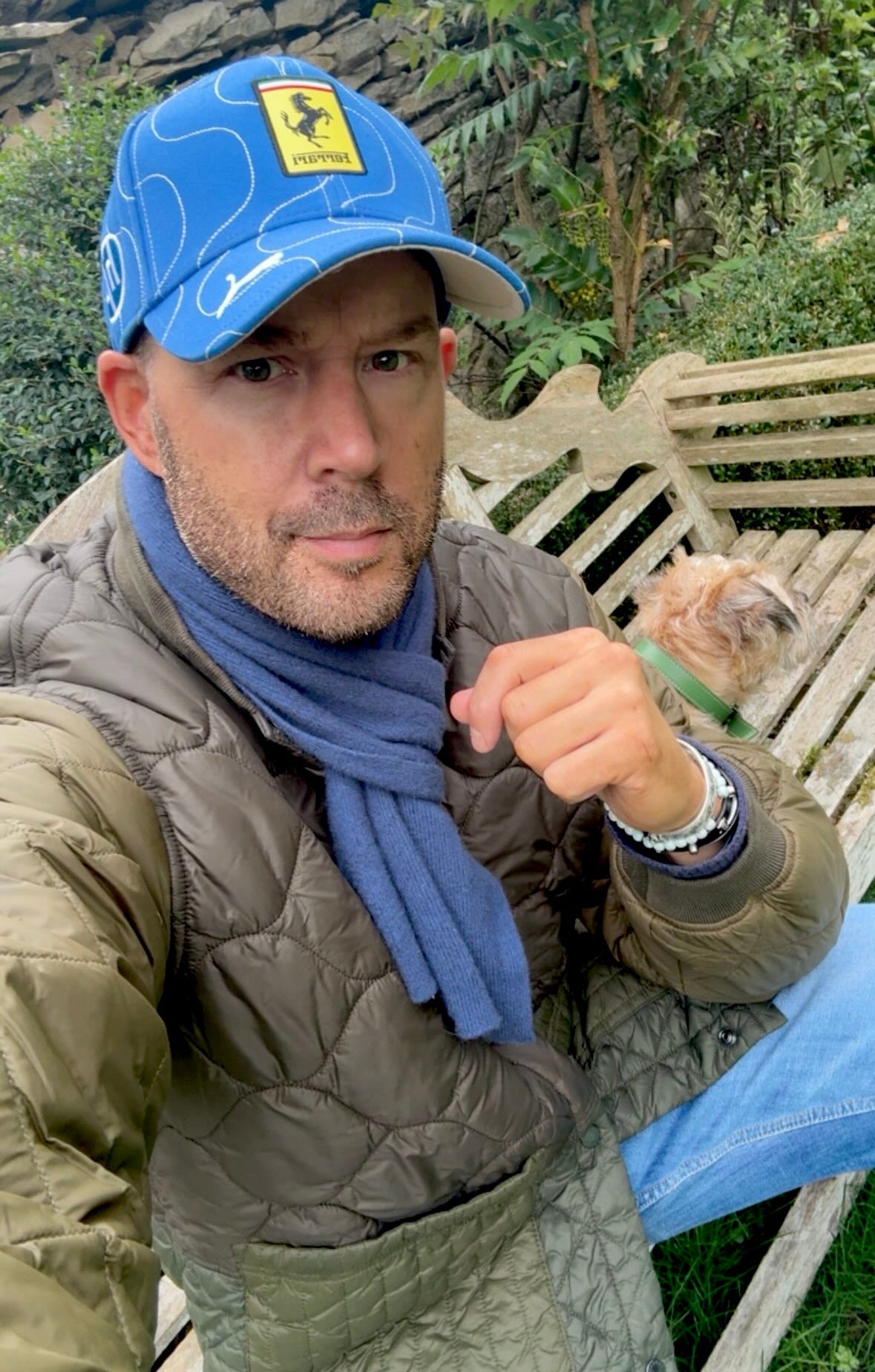 Artist Teddy McDonald wearing a blue Ferrari cap, Barbour quilted jacket and blue scarf, sitting outdoors with a Border Terrier dog on a bench in The Cotswolds garden.